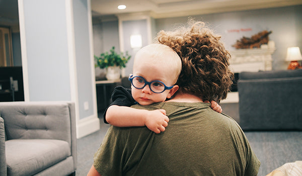 A parent carries her baby over their shoulder. The baby is wearing glasses and stairing off into the distance.