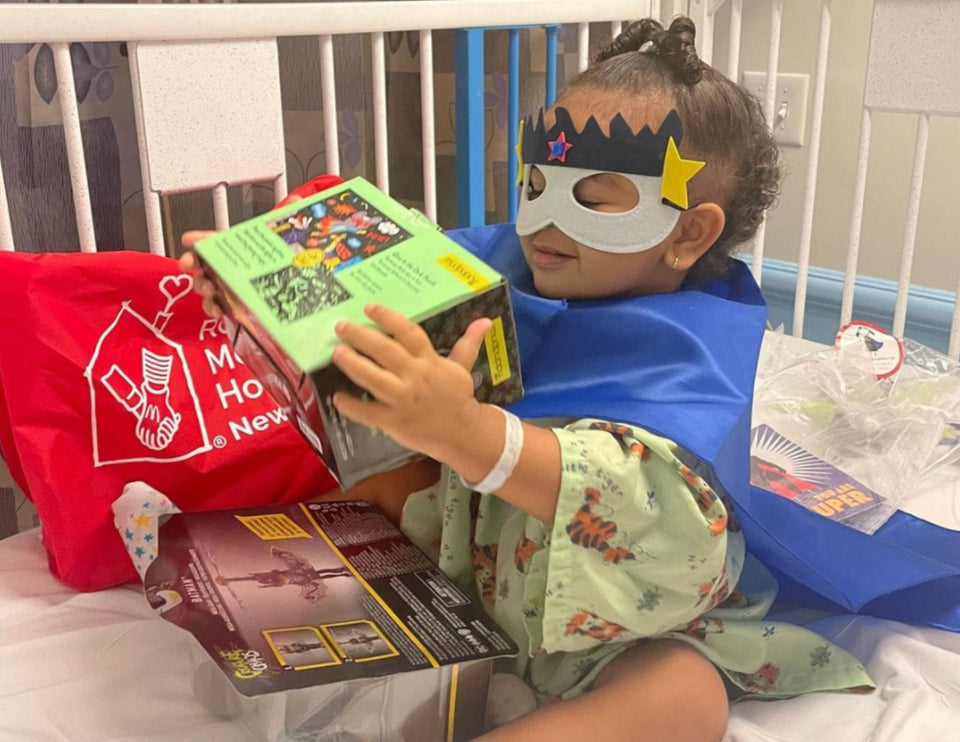 A superhero child holds up a gift she just got from Ronald McDonald House New York. She wears a cape and a mask in her hospital bed.