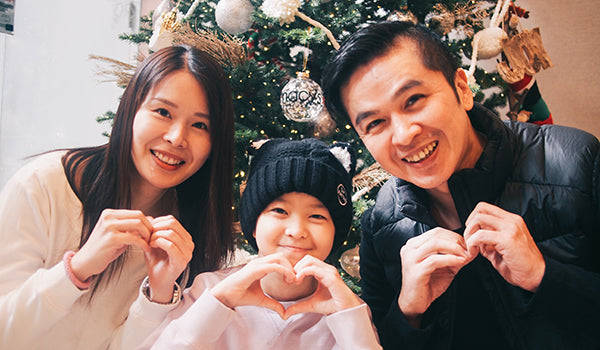 A family of three pose in front of a tree at the Ronald McDonald House New York. They make heart symbols with their hands and are smiling for the camera. From right to left: Mother, daughter, father.