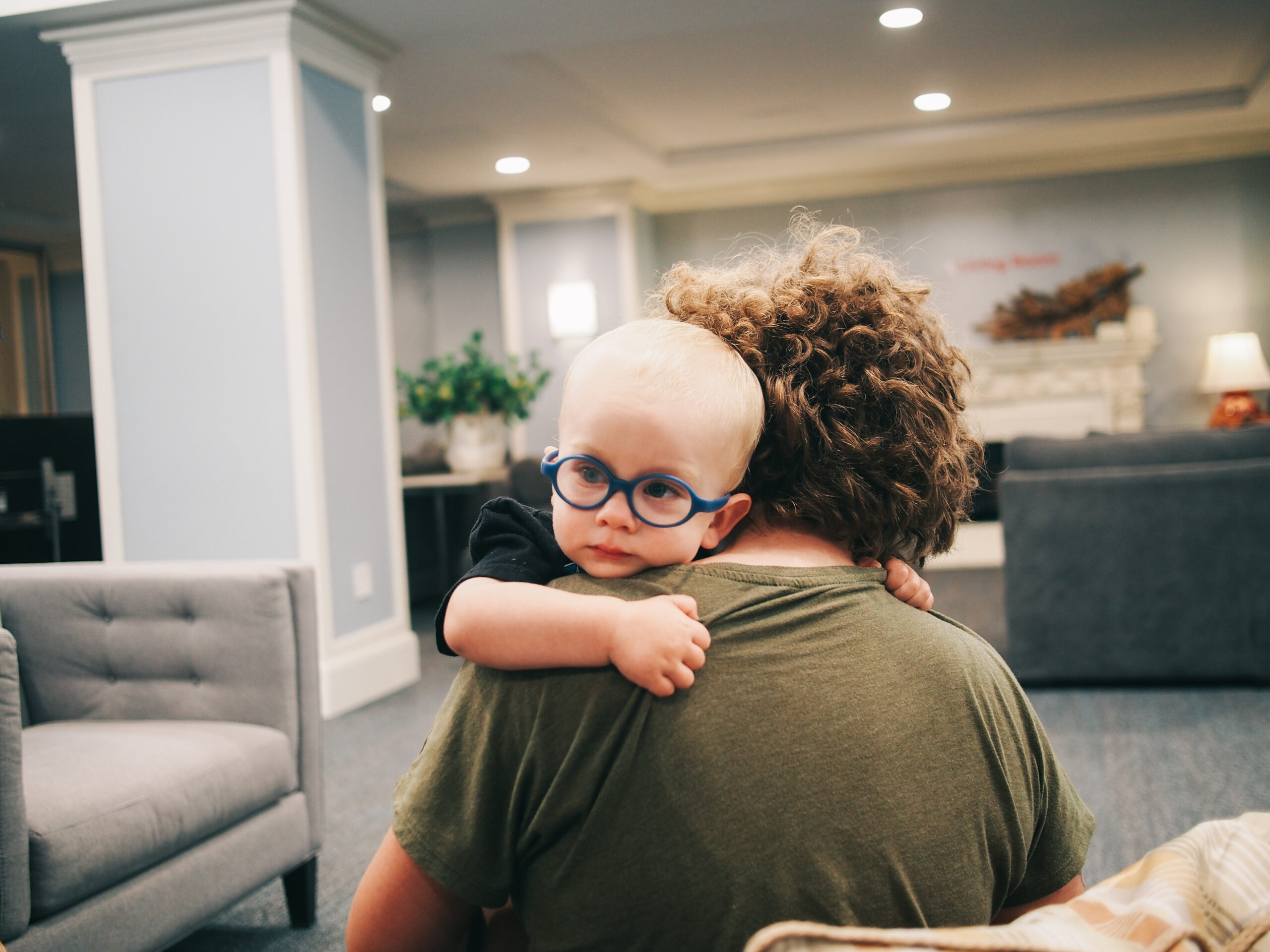 A little boy with glasses is being held over his mother's shoulder. Her back is turned to the camera, but he is looking off into the distance in the direction of the camera.